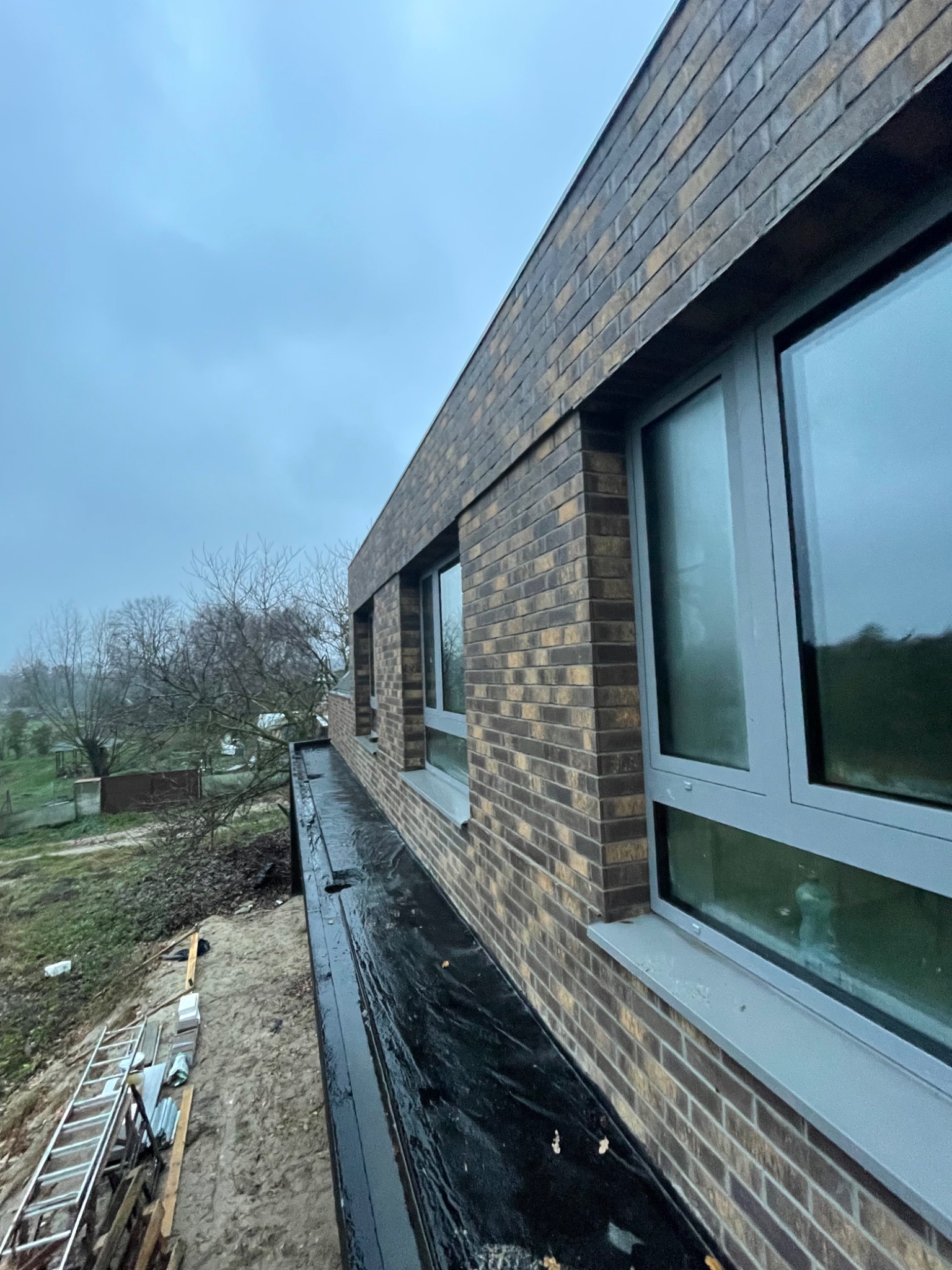 Fragment nowo wybudowanego domu z brązowej cegły z szarymi oknami, widok z góry na dach i otaczający teren w pochmurny dzień.