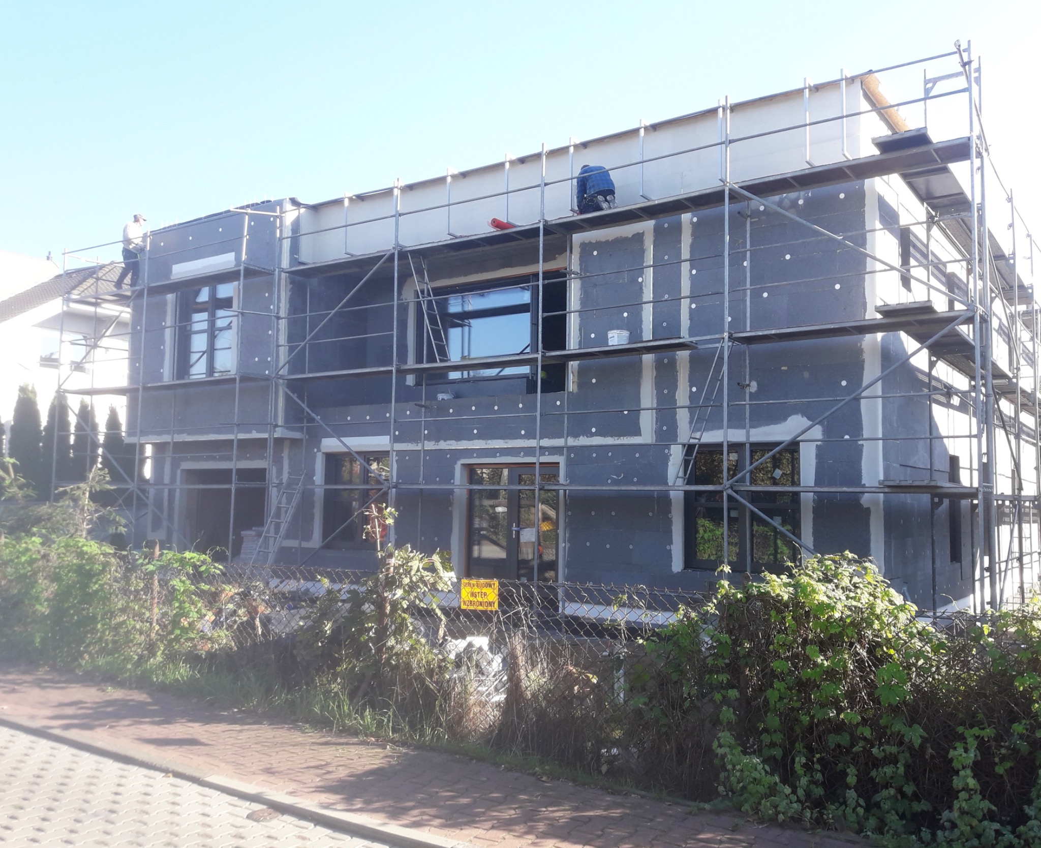 Dwukondygnacyjny budynek w trakcie ocieplania elewacji, z widocznymi rusztowaniami, częściowo otynkowany, z zamontowanymi oknami, na pierwszym planie ogrodzenie i zieleń.