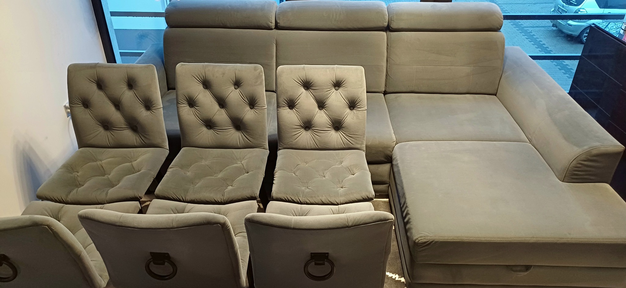 Szary narożnik z podnoszonymi zagłówkami i krzesła tapicerowane z guzikami, ustawione w salonie meblowym.