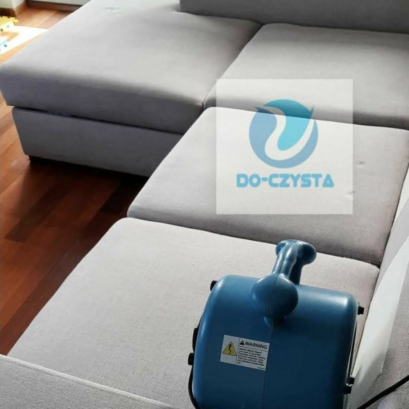 Szara, modułowa kanapa narożna z widocznym niebieskim urządzeniem do czyszczenia tapicerki, częściowo zasłonięta naklejką z logiem firmy 'DO-CZYSTA'.