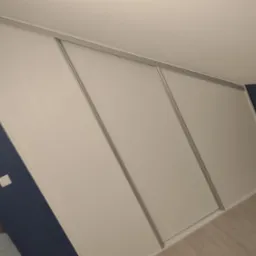 Biała szafa wnękowa z przesuwnymi drzwiami zamontowana na poddaszu o skośnych ścianach, widoczna na tle granatowej ściany.