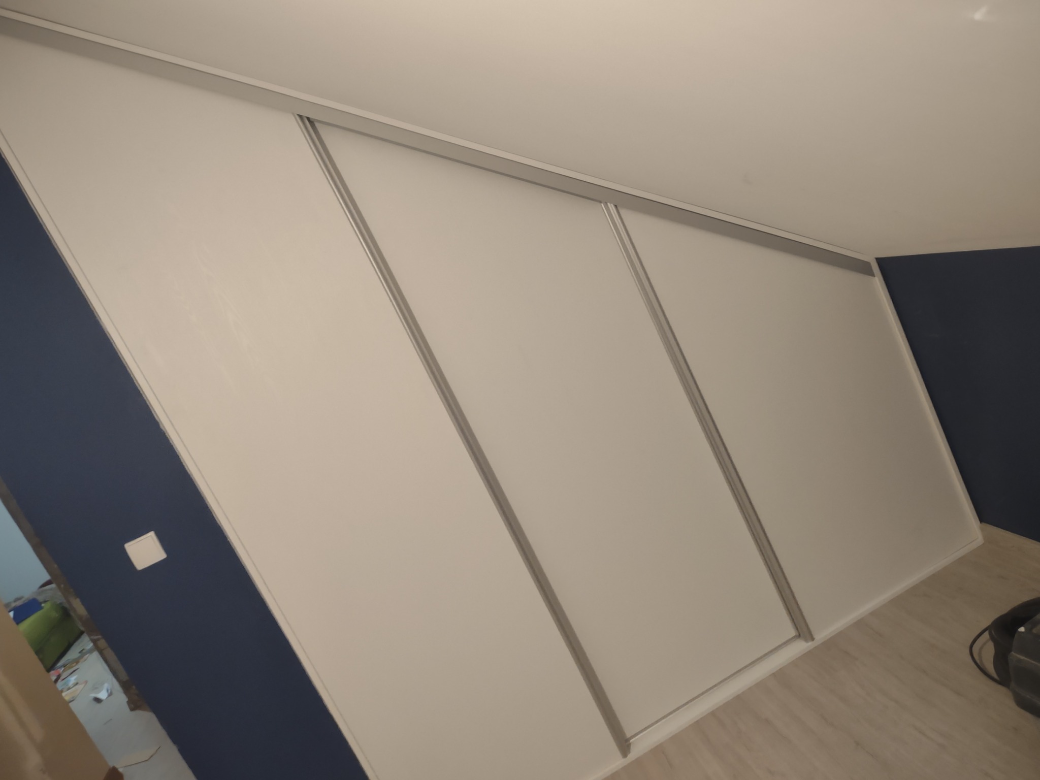 Biała szafa wnękowa z przesuwnymi drzwiami zamontowana na poddaszu o skośnych ścianach, widoczna na tle granatowej ściany.