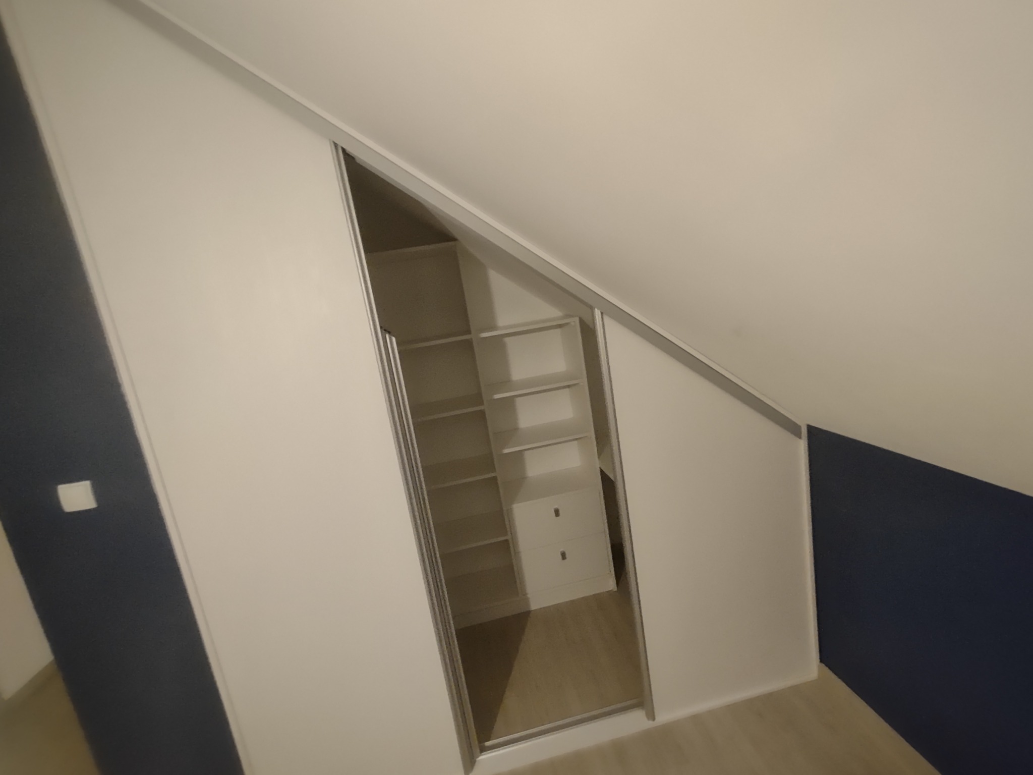 Biała szafa wnękowa z przesuwnymi drzwiami, wkomponowana w skos dachu na poddaszu, widoczne półki i szuflady wewnątrz.