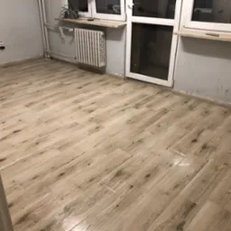 Ułożone panele podłogowe w jasnym odcieniu drewna, widoczne grzejniki i fragment okna z parapetem, ściany w odcieniach szarości, widok z perspektywy wejścia do pomieszczenia.