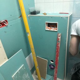 VerdeBud - Montaż płyty gipsowo-kartonowej w łazience z widocznymi poziomnicami, wiertarką i puszką elektryczną.