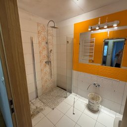 VerdeBud - Remont łazienki: kabina prysznicowa z wzorem kwiatowym, pomarańczowe płytki lustrzane, białe płytki, wiadro i częściowo zdemontowana armatura. Widoczne ślady prac remontowych.