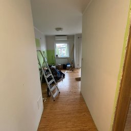 VerdeBud - Korytarz w trakcie malowania: ściany w jasnym odcieniu, widoczna drabina, narzędzia malarskie i fragment podłogi z paneli. W tle okno z widokiem na budynek.