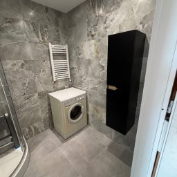 VerdeBud - Wykończona łazienka z marmurowymi płytkami, pralką, grzejnikiem drabinkowym i czarną szafką wiszącą. Nowoczesny design z szarymi płytkami podłogowymi.