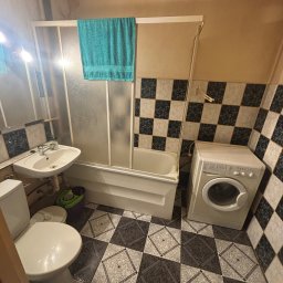 VerdeBud - Ukończona łazienka z wanną za parawanem, umywalką, toaletą i pralką. Czarno-białe płytki na podłodze i ścianach. Turkusowy ręcznik na parawanie.