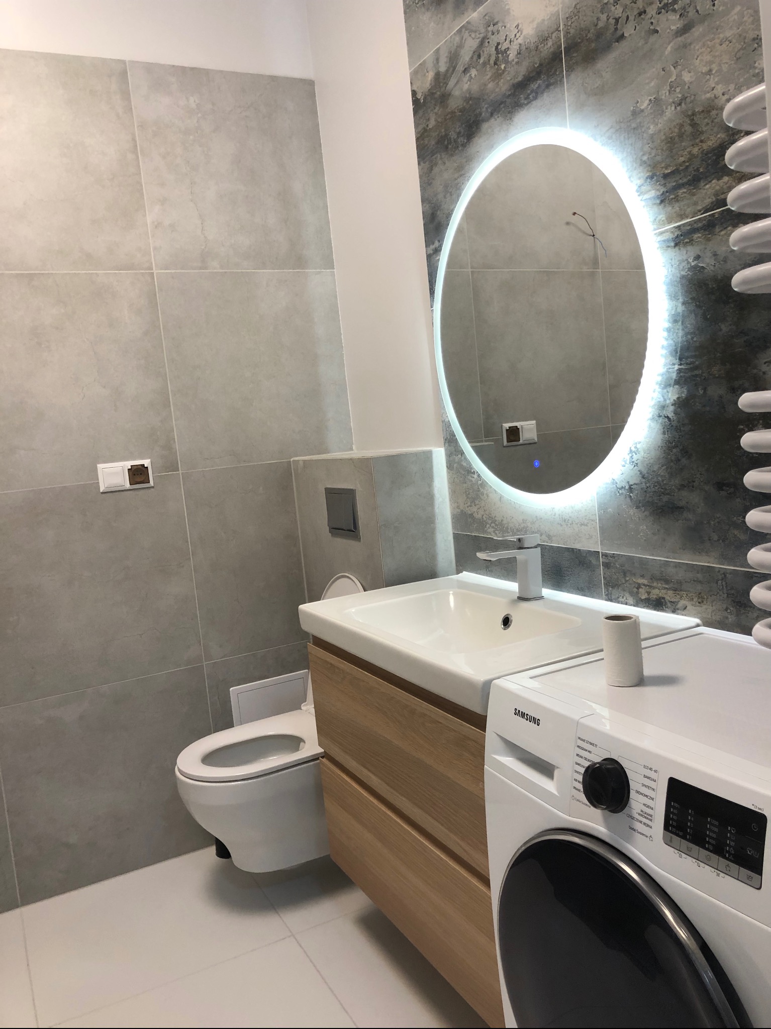 Nowoczesna łazienka z szarymi płytkami, owalnym lustrem z podświetleniem LED, drewnianą szafką pod umywalką, toaletą w zabudowie i pralką Samsung.