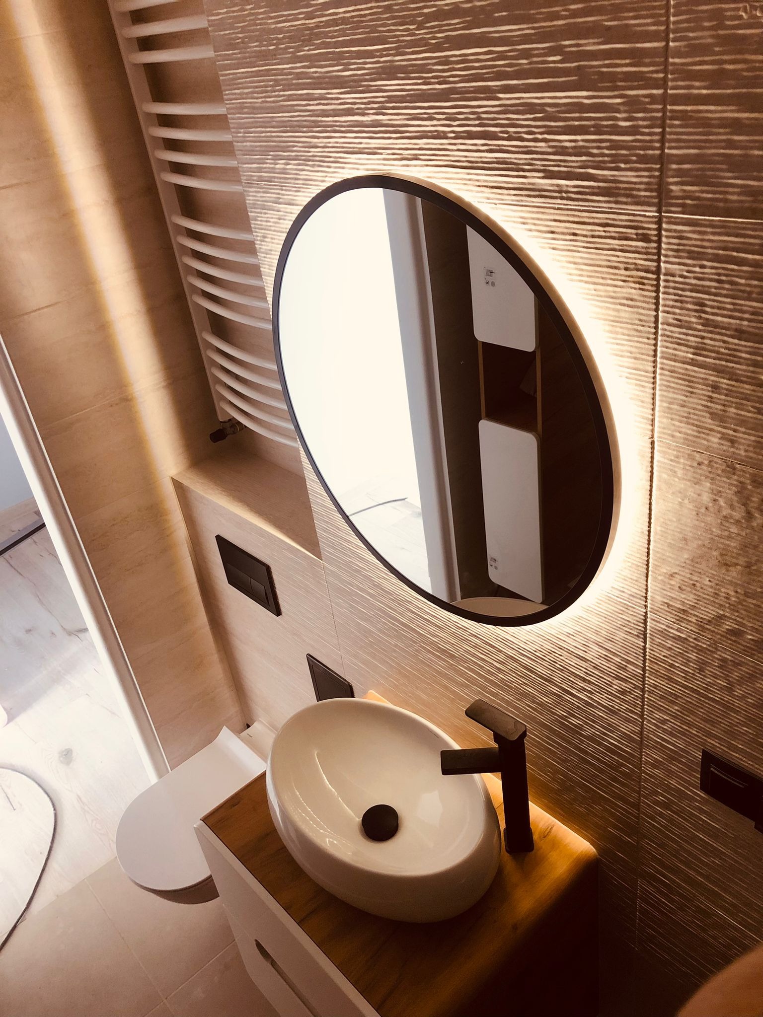 Nowoczesna łazienka z owalnym lustrem podświetlanym LED, umywalką nablatową na drewnianym blacie i teksturowanymi płytkami ściennymi.