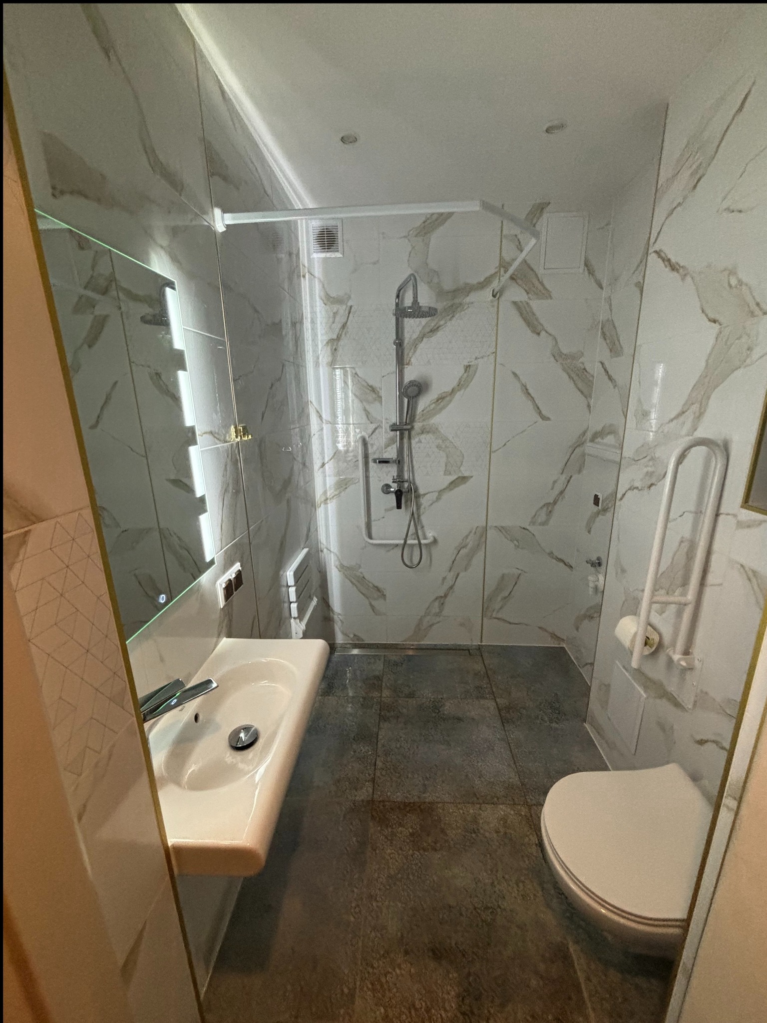 Nowoczesna łazienka z biało-szarymi marmurkowymi płytkami, podświetlanym lustrem nad umywalką, kabiną prysznicową z deszczownicą i uchwytami dla osób niepełnosprawnych przy toalecie.