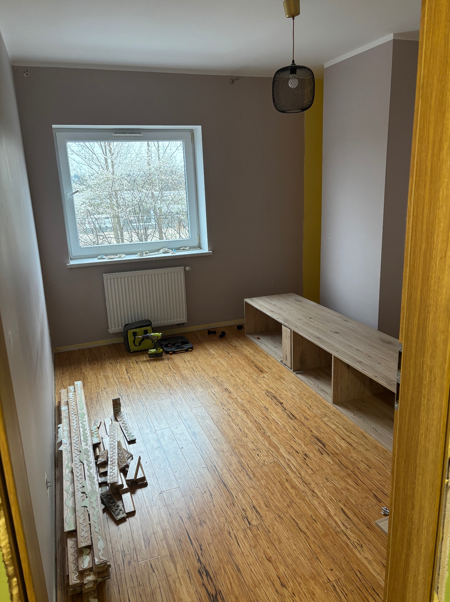 Pokój po remoncie: ułożone panele podłogowe, listwy przypodłogowe przy ścianie, zamontowany regał, okno z grzejnikiem, narzędzia pracy. Widok zza otwartych drzwi.