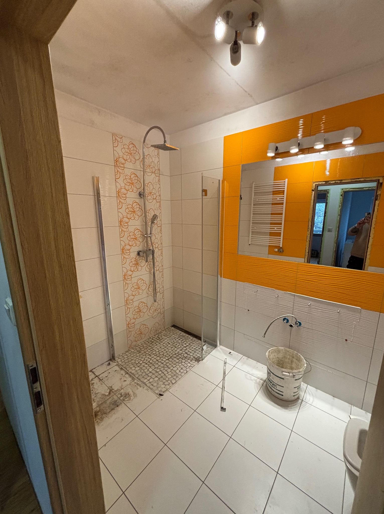 Remont łazienki: kabina prysznicowa z wzorem kwiatowym, pomarańczowe płytki lustrzane, białe płytki, wiadro i częściowo zdemontowana armatura. Widoczne ślady prac remontowych.