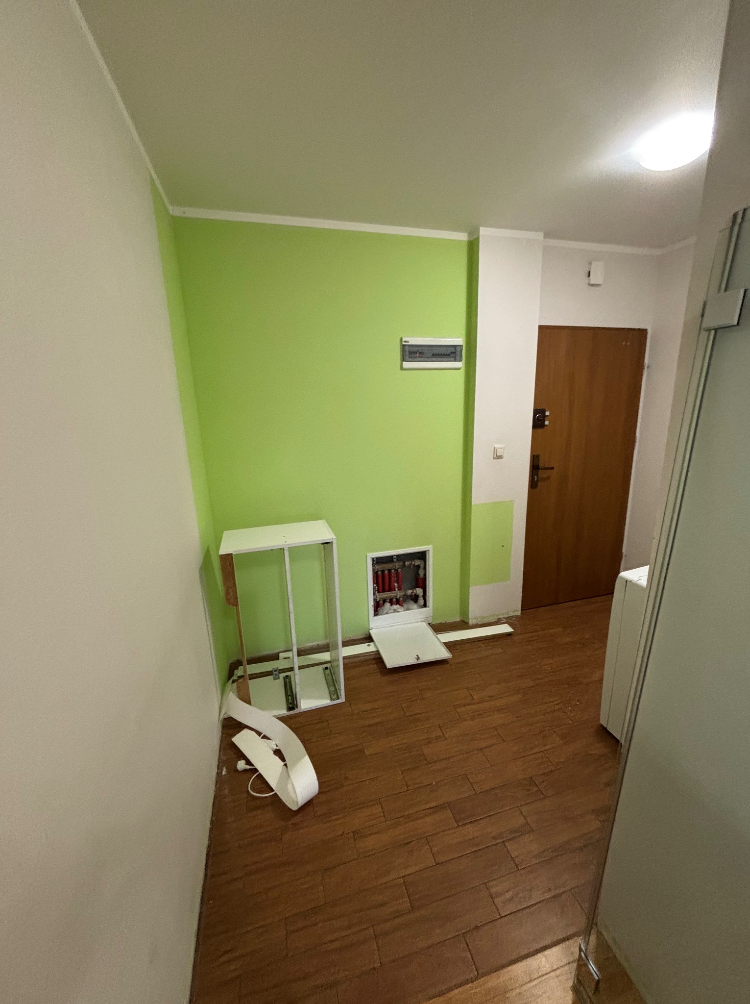 Wnętrze w trakcie remontu: zielona ściana, brązowe panele podłogowe, otwarta skrzynka z instalacją elektryczną, fragmenty mebli do montażu. Widok na drzwi wejściowe.