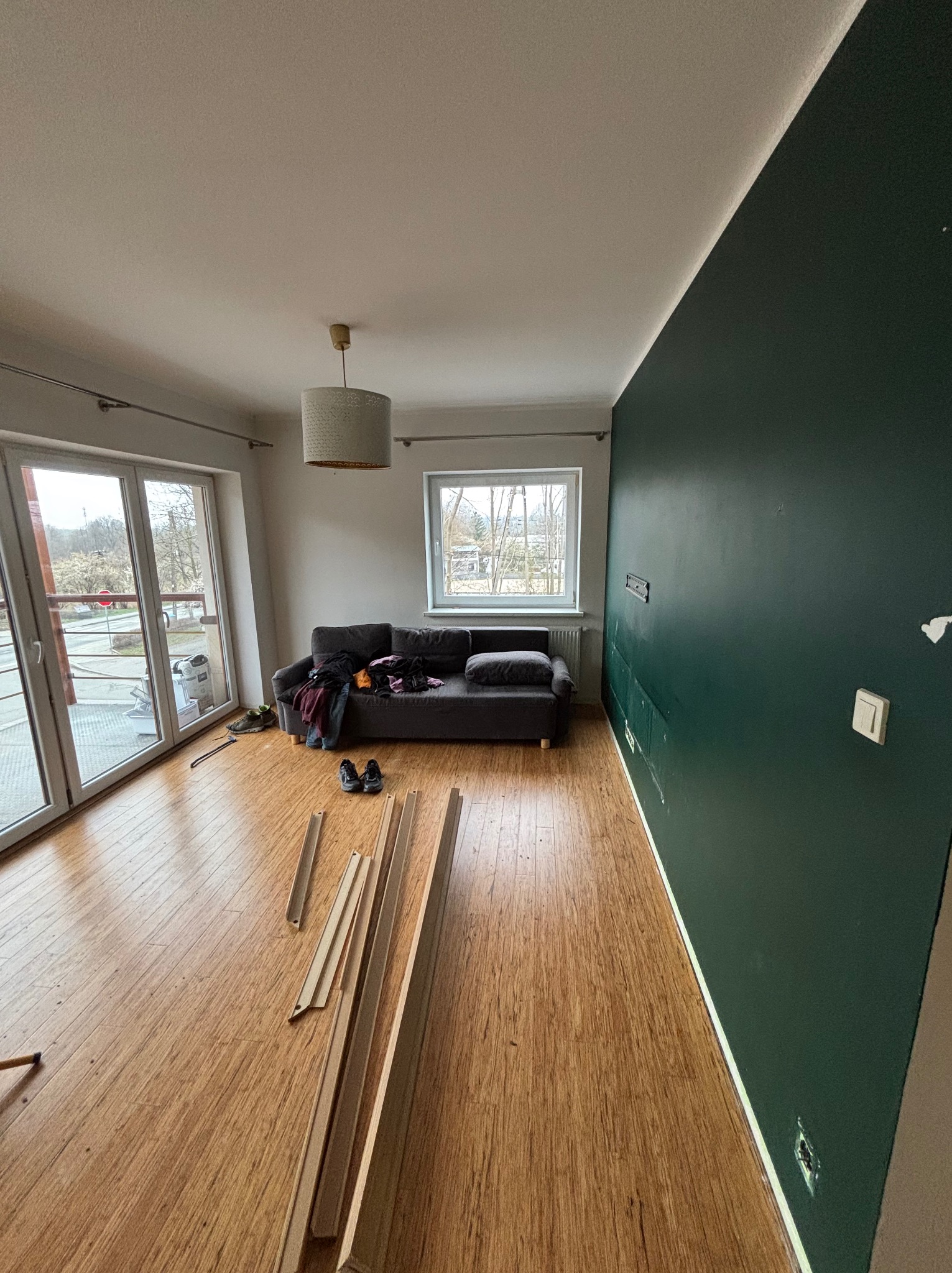 Pokój przed wykończeniem: panele podłogowe, pomalowana na ciemnozielono ściana, okno i balkon. Widoczne listwy przypodłogowe i sofa.