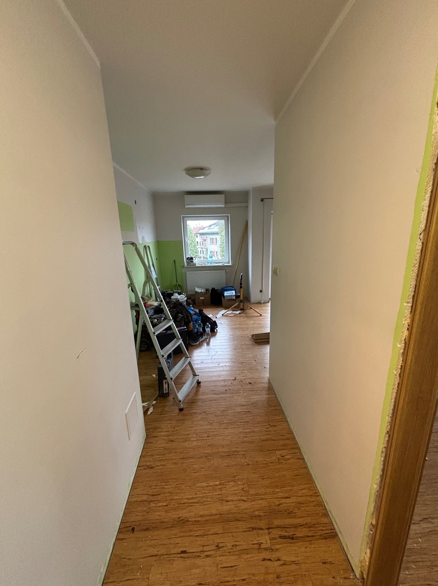 Korytarz w trakcie malowania: ściany w jasnym odcieniu, widoczna drabina, narzędzia malarskie i fragment podłogi z paneli. W tle okno z widokiem na budynek.