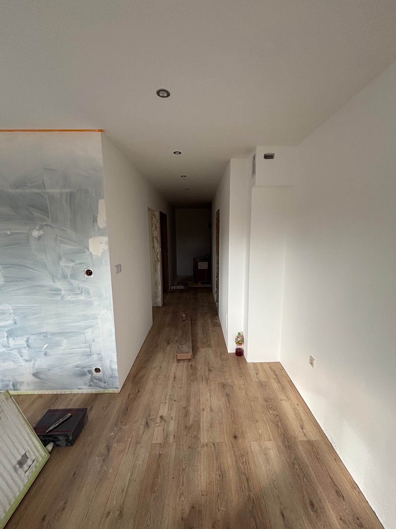 Korytarz w trakcie remontu: świeżo ułożone panele podłogowe, ściany przygotowane pod malowanie, widoczne narzędzia glazurnicze. W tle widoczne kolejne pomieszczenia.