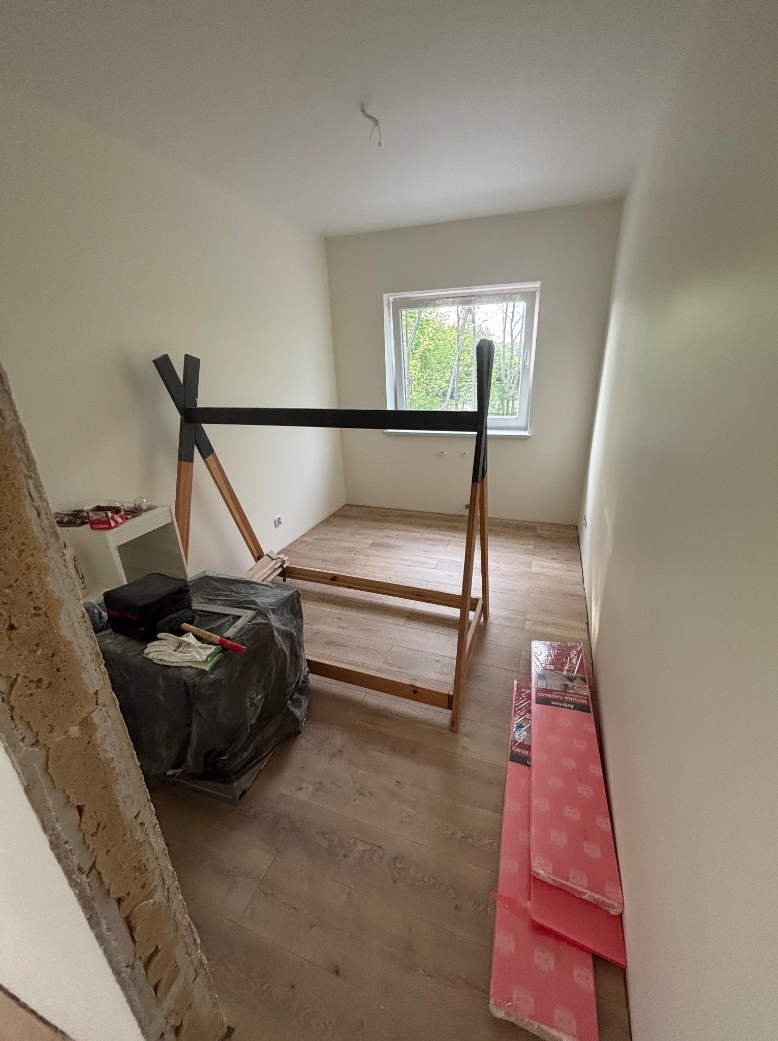 Pokój po remoncie: drewniana podłoga, okno z widokiem na zieleń, fragment konstrukcji łóżka i narzędzia budowlane. Widoczny etap wykończenia wnętrza.