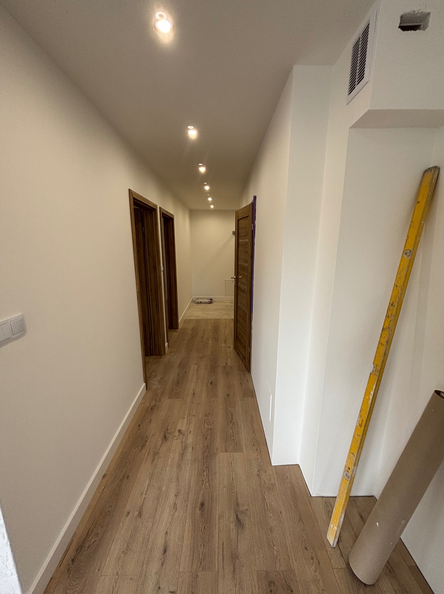 Korytarz po remoncie z panelami podłogowymi, świeżo pomalowanymi ścianami i zamontowanymi drzwiami. Poziomica i rolka papieru przy ścianie.