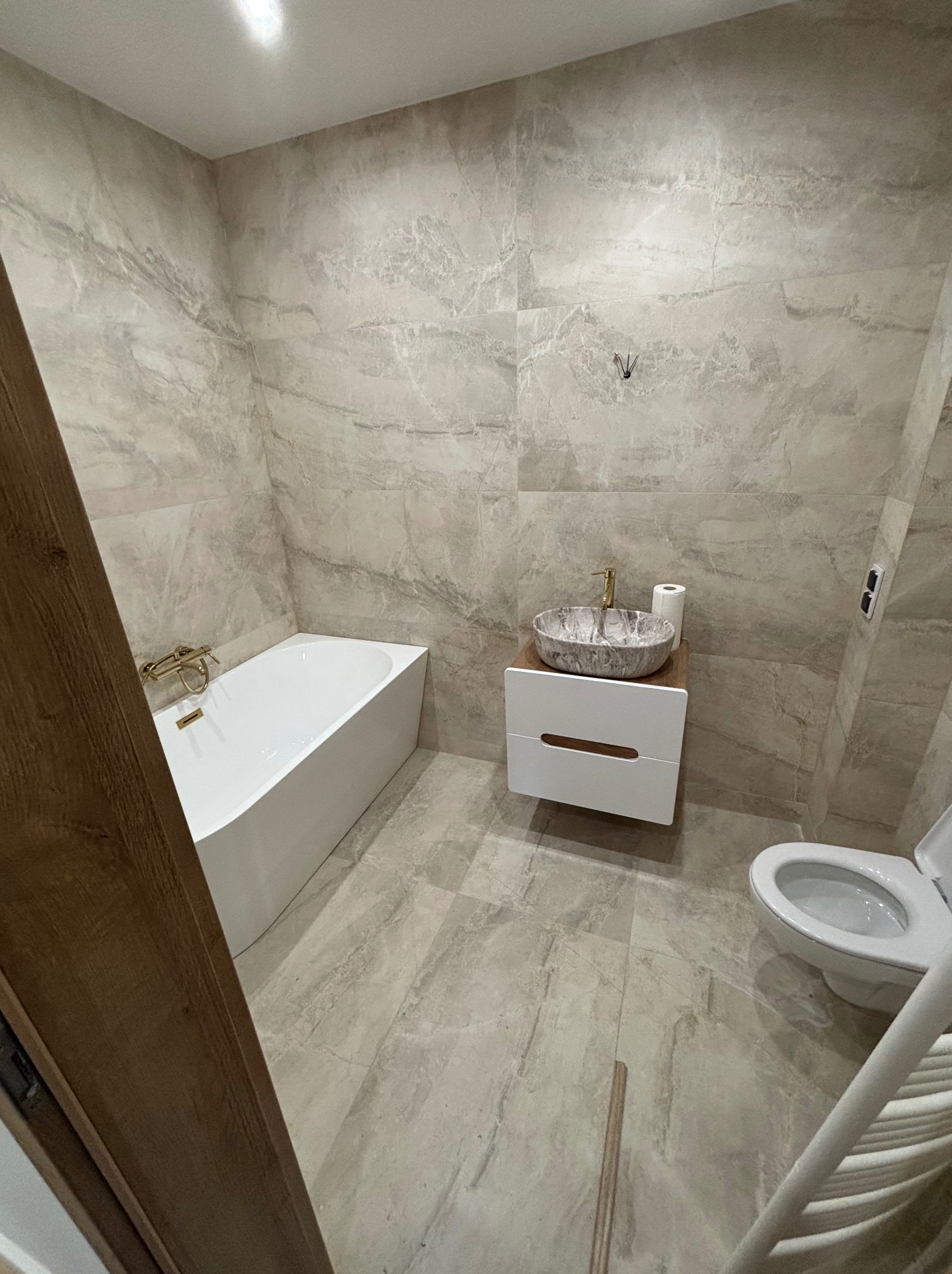 Wykończona łazienka w beżowych płytkach imitujących marmur. Umywalka nablatowa w odcieniach szarości, biała wanna i toaleta.