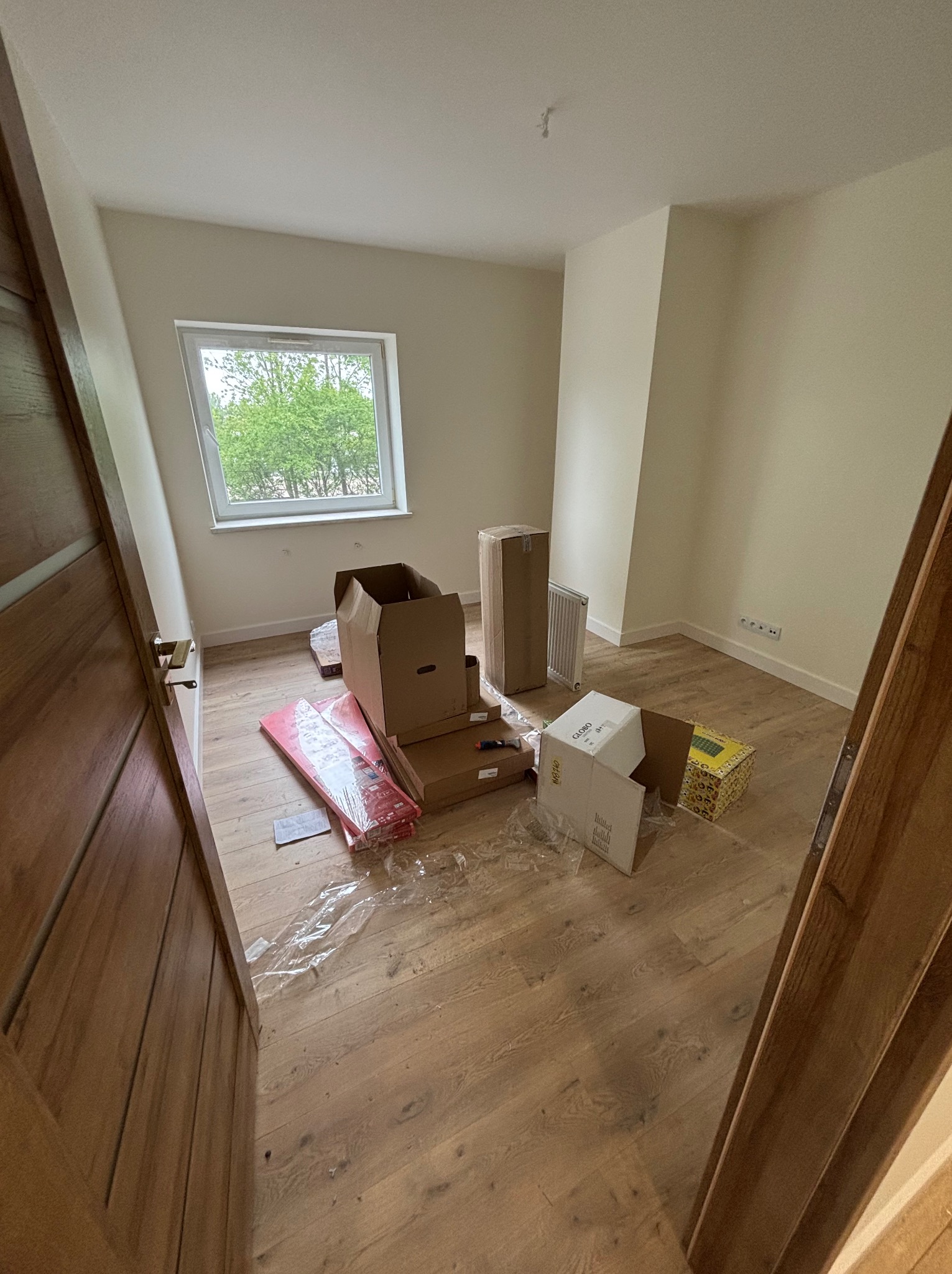 Puste pomieszczenie z oknem, na podłodze panele i kartony. Widoczny fragment drewnianych drzwi z dekoracyjnymi listwami i klamką. Przygotowania do wykończenia.