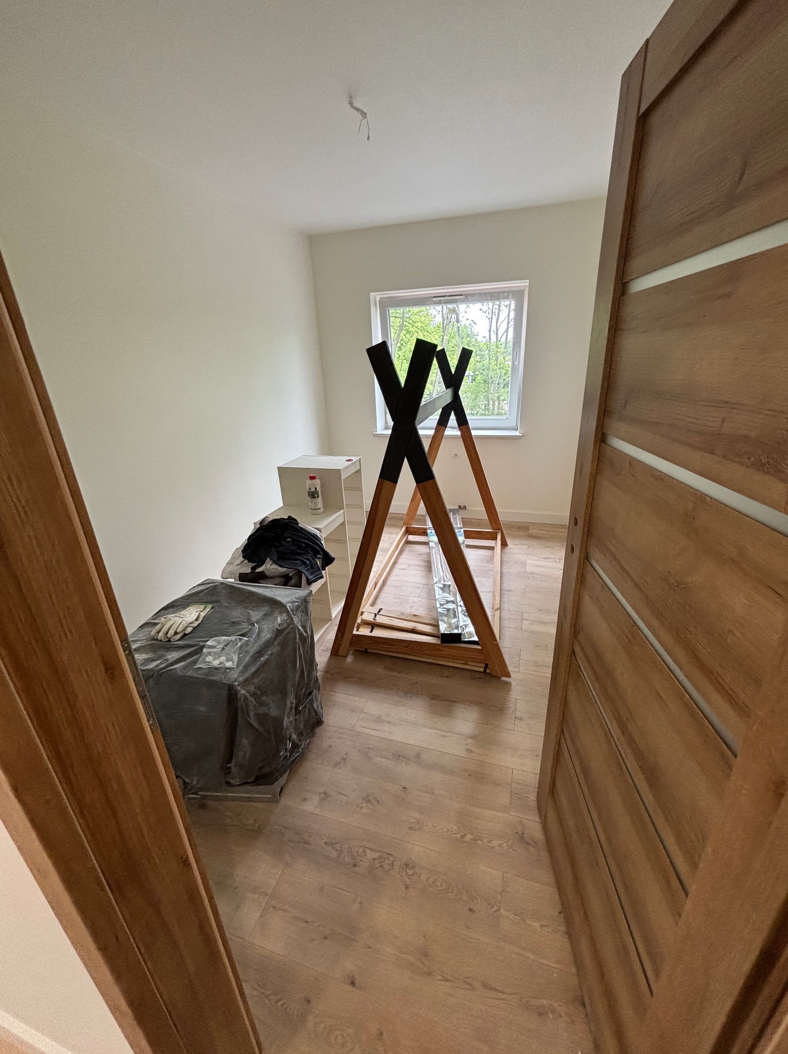 Pokój z drewnianą podłogą i ramą tipi pod oknem, widok przez otwarte, drewniane drzwi. Częściowo zakryte meble i akcesoria remontowe.
