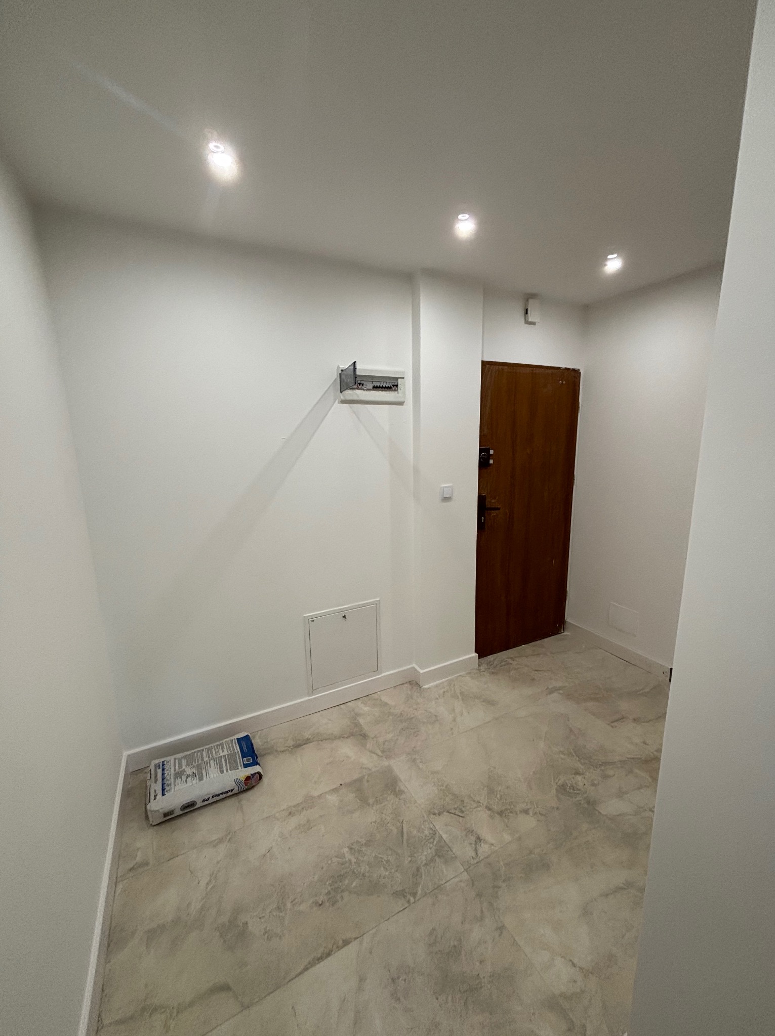 Wykończony przedpokój z marmurową podłogą i białymi ścianami. Widoczna torba z klejem do płytek przy ścianie, brązowe drzwi i skrzynka elektryczna.