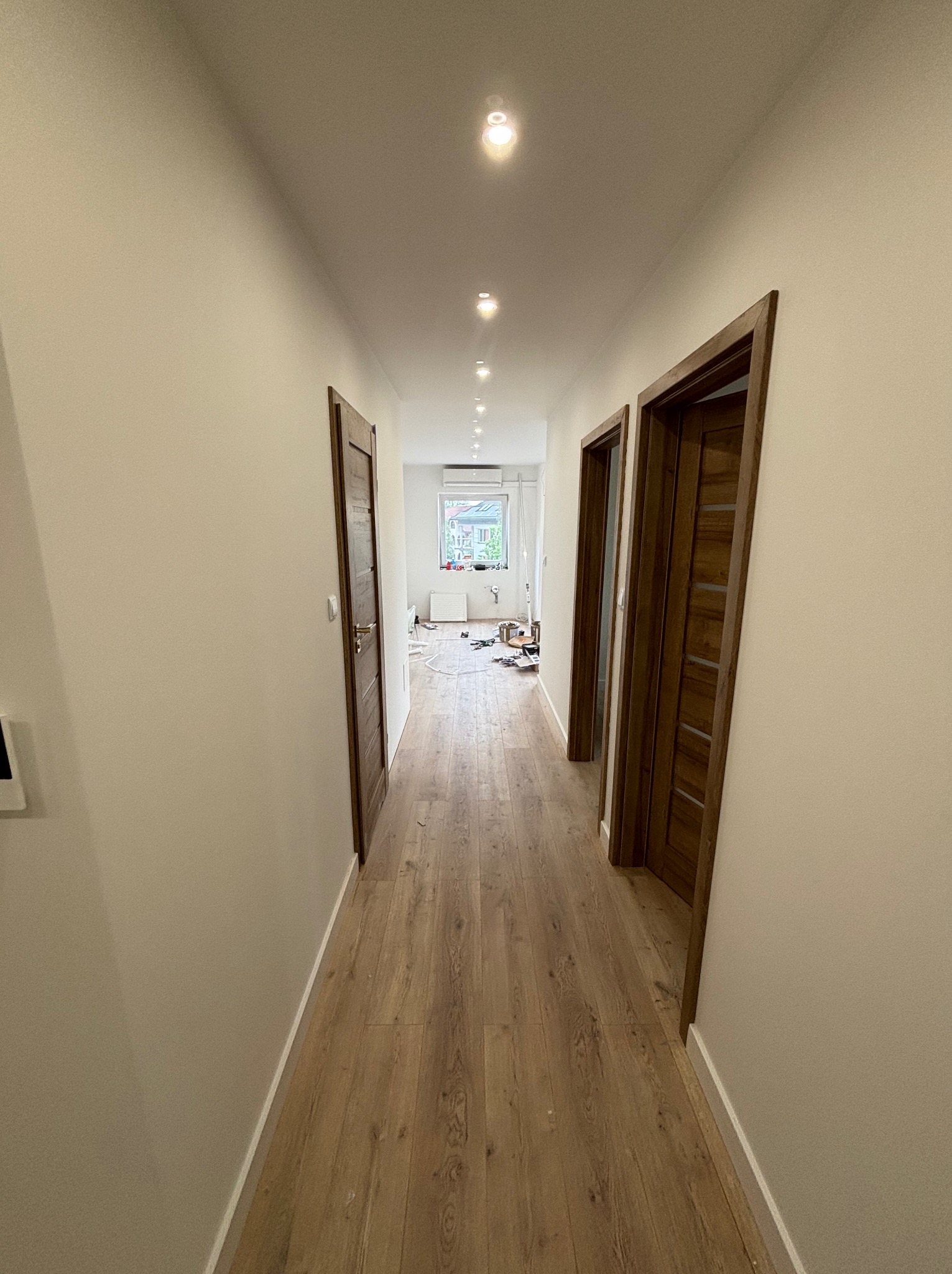 Wykończony korytarz z drewnianą podłogą i dwiema parami drewnianych drzwi. Widoczne punktowe oświetlenie sufitowe i fragment pomieszczenia z oknem w tle.