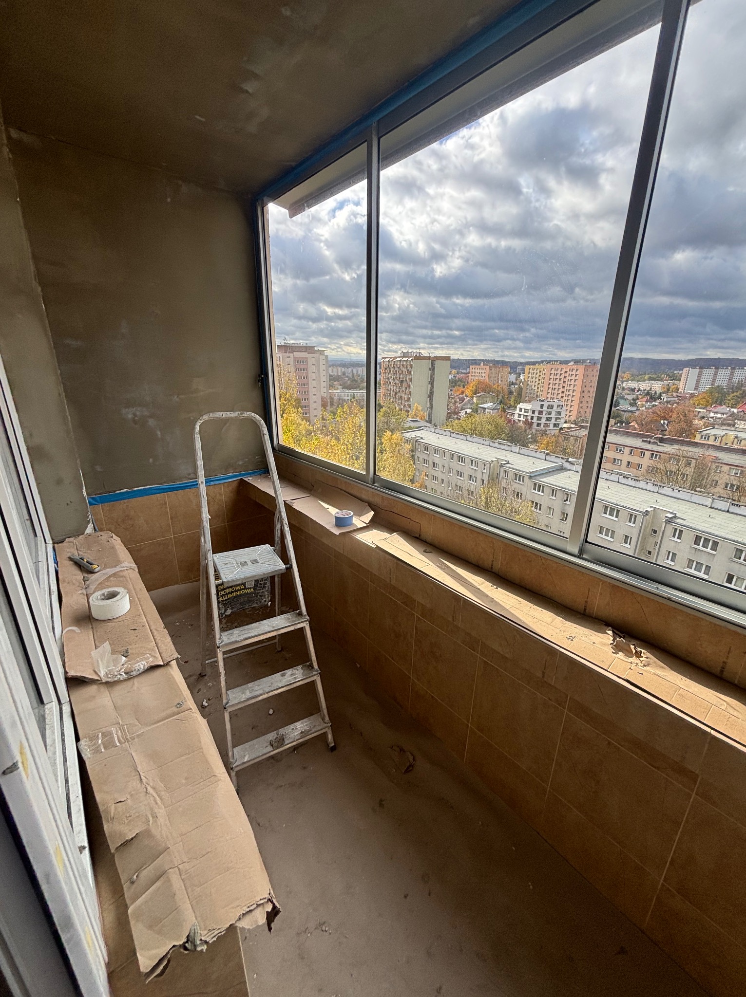 Balkon w trakcie prac wykończeniowych: ściany po gładzi, okna zamontowane, widoczna drabina i materiały zabezpieczające. Panorama miasta w tle.