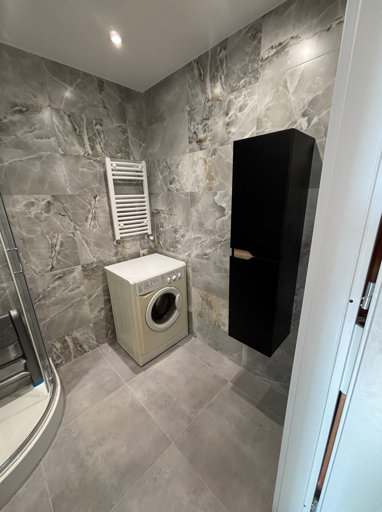 Wykończona łazienka z marmurowymi płytkami, pralką, grzejnikiem drabinkowym i czarną szafką wiszącą. Nowoczesny design z szarymi płytkami podłogowymi.
