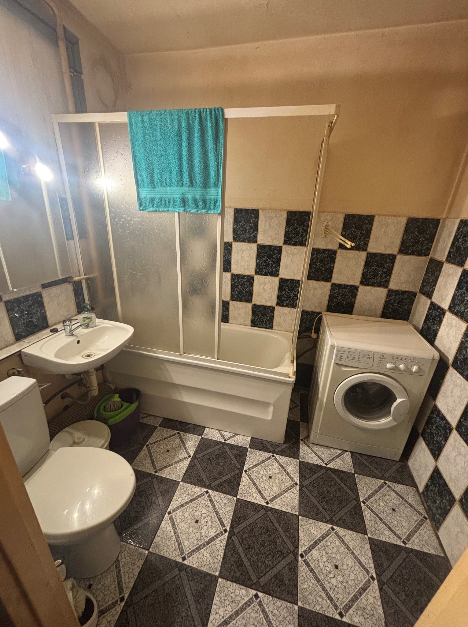 Ukończona łazienka z wanną za parawanem, umywalką, toaletą i pralką. Czarno-białe płytki na podłodze i ścianach. Turkusowy ręcznik na parawanie.