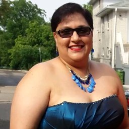 Rodriguez andre - Uśmiechnięta kobieta w niebieskiej sukni bez ramiączek i dużym niebieskim naszyjniku, stojąca na zewnątrz, z budynkiem i zielenią w tle.