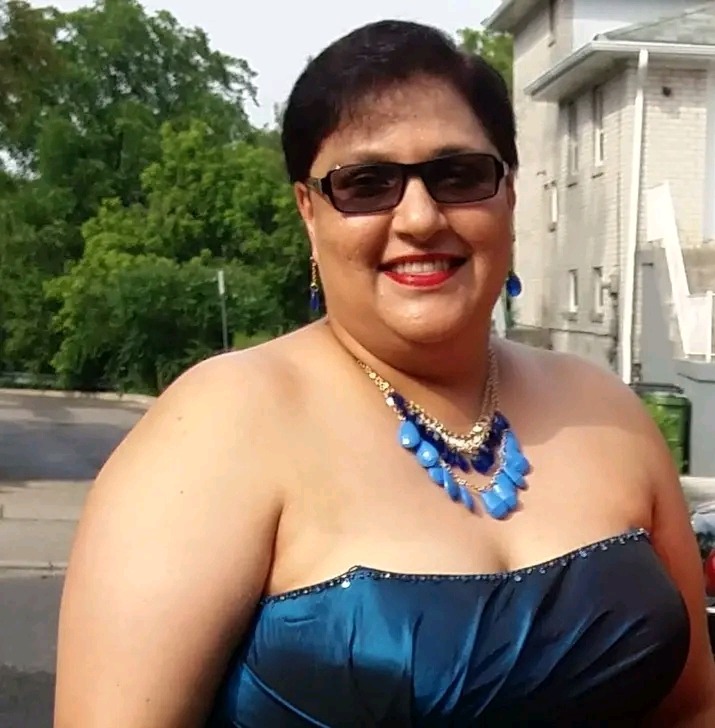 Uśmiechnięta kobieta w niebieskiej sukni bez ramiączek i dużym niebieskim naszyjniku, stojąca na zewnątrz, z budynkiem i zielenią w tle.