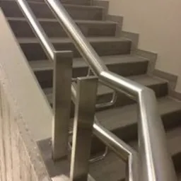 Schody wyłożone szarymi płytkami z metalową, nierdzewną barierką, widok z dołu ukazujący konstrukcję mocowania poręczy do stopni.