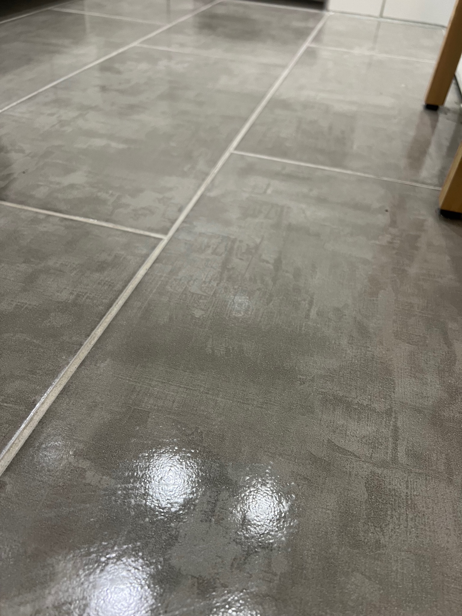 Szare, matowe płytki podłogowe z jasnymi fugami, częściowo odbijające światło, widoczne nogi drewnianego krzesła w prawym górnym rogu.