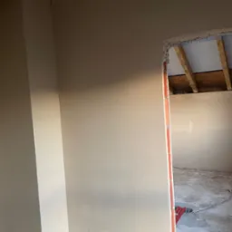 Gładka, świeżo nałożona warstwa tynku na ścianie wewnętrznej, widoczne surowe wykończenie nadproża drzwiowego i fragment nieotynkowanego sufitu z drewnianą konstrukcją.