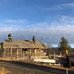 K-Maro - Budowa domu z zielonym dachem: dwóch pracowników w żółtych kamizelkach montuje konstrukcję dachu, widoczna przyczepa z napisem 'GAUPEN' i krajobraz górski w tle.