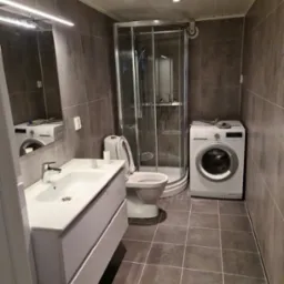 Szara łazienka z kabiną prysznicową, umywalką z szafką i pralką, widok z niskiego kąta, z otwartą klapą toalety.