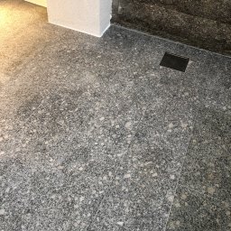 K-Maro - Szara, nakrapiana podłoga z płytek ceramicznych, z odpływem liniowym, przylegająca do ściany wyłożonej kamieniem dekoracyjnym i wykończonego narożnika ściany.