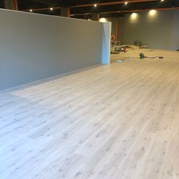 K-Maro - Jasne panele podłogowe ułożone w nowym pomieszczeniu, widoczne narzędzia i materiały budowlane w tle, ściany w odcieniach szarości, czarne oświetlenie sufitowe.