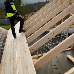 K-Maro - Pracownik w odblaskowej kurtce stoi na drewnianej konstrukcji dachu w trakcie budowy, w tle widok na zalesione wzgórza i pochmurne niebo.