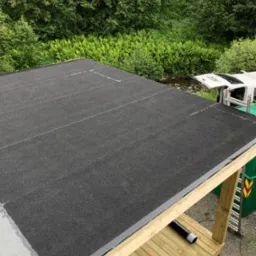 Czarna papa na dachu z widocznymi liniami łączeń, Koszalin, w tle bujna roślinność i częściowo widoczny samochód dostawczy.