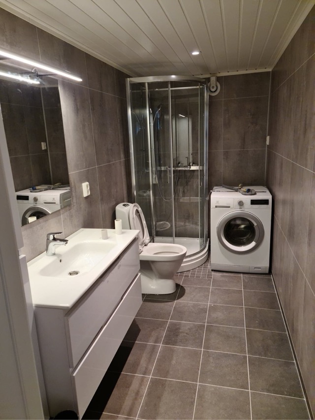 Szara łazienka z kabiną prysznicową, umywalką z szafką i pralką, widok z niskiego kąta, z otwartą klapą toalety.