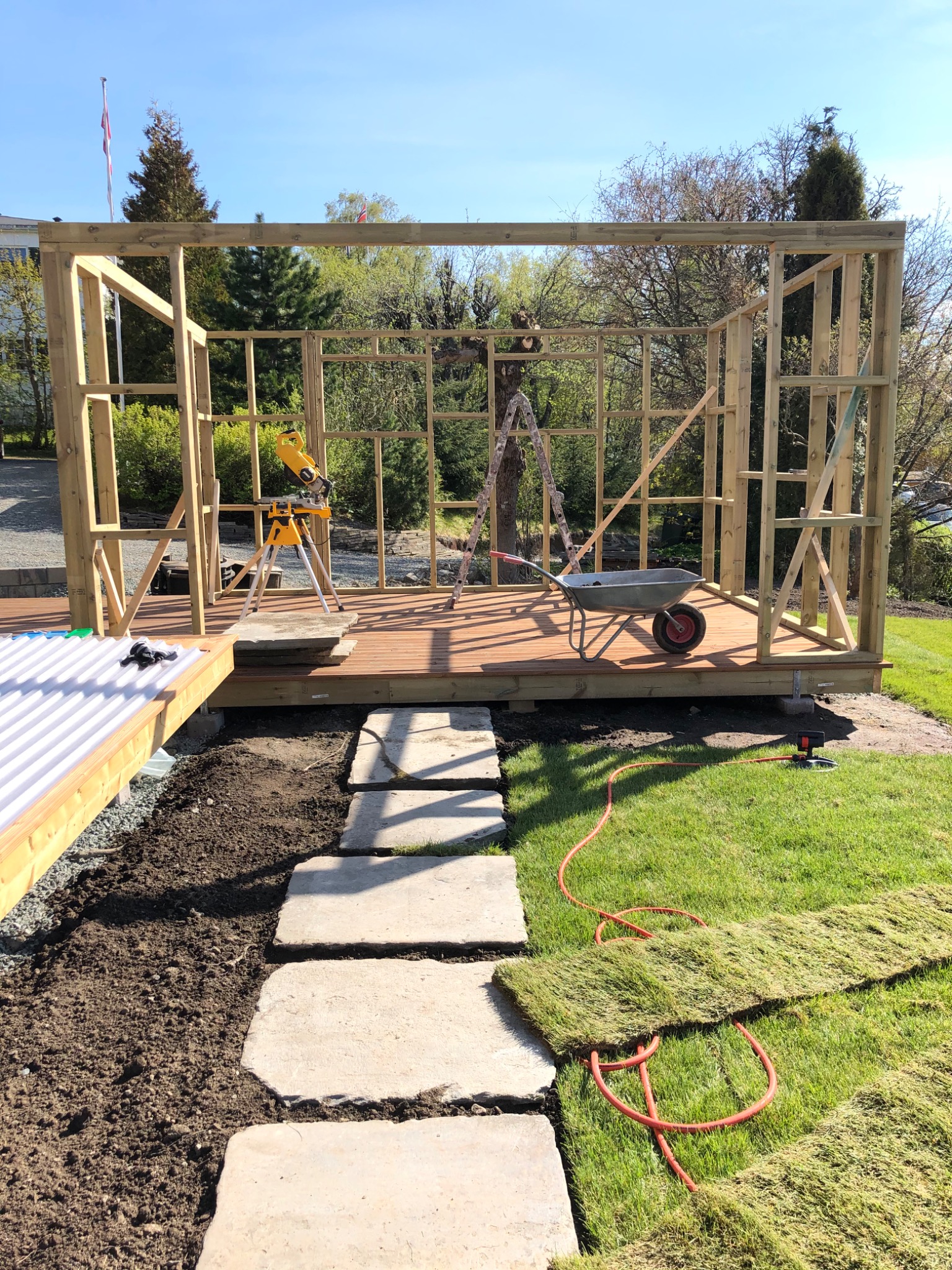 Konstrukcja szkieletowa drewnianej altany w trakcie budowy, widoczna taczka, piła stołowa i rozłożona darń na trawniku.