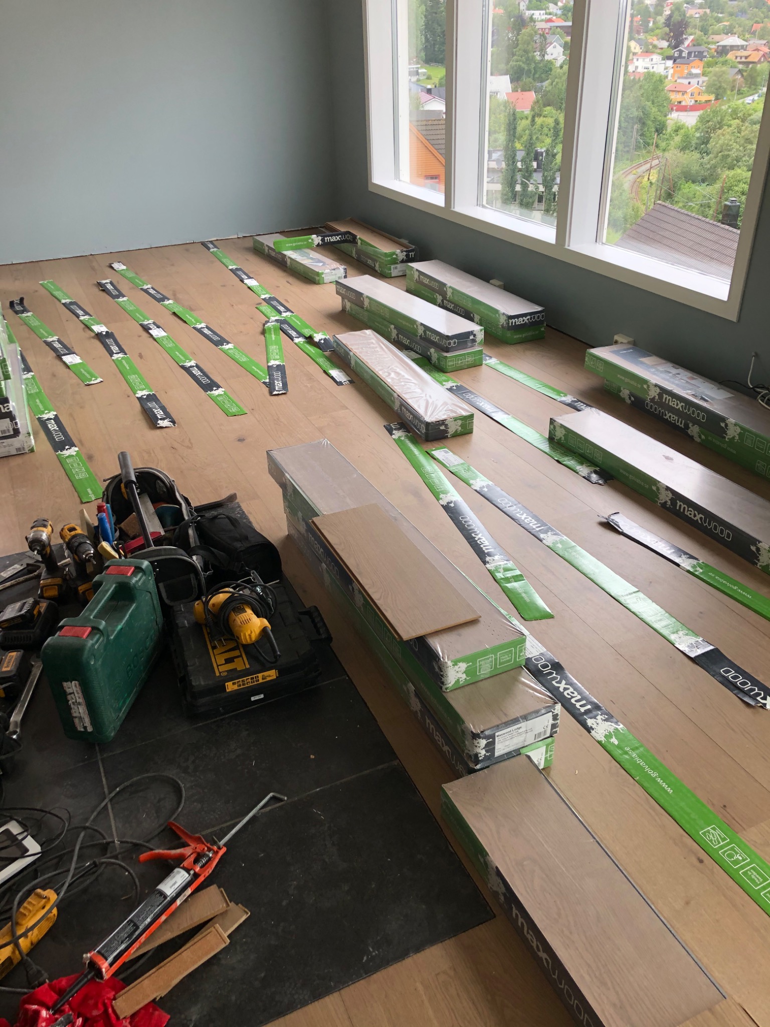 Układanie paneli Maxwood Lodge: widok na rozpakowane paczki paneli, narzędzia i listwy podłogowe w jasnym pomieszczeniu z dużymi oknami.