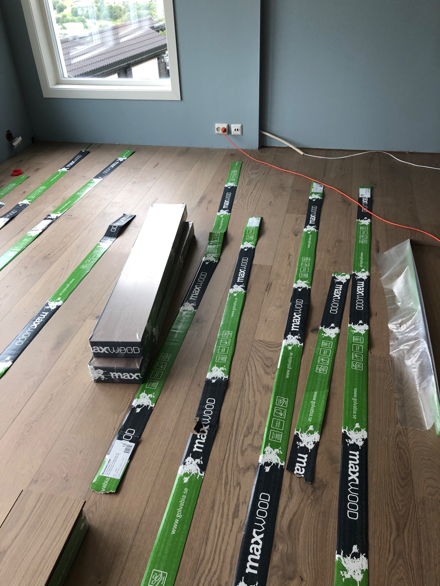 Przygotowanie do ułożenia paneli podłogowych marki Maxwood, widoczne paczki paneli i listwy opakowaniowe z logo na drewnianej podłodze w pomieszczeniu z błękitnymi ścianami i oknem dachowym.