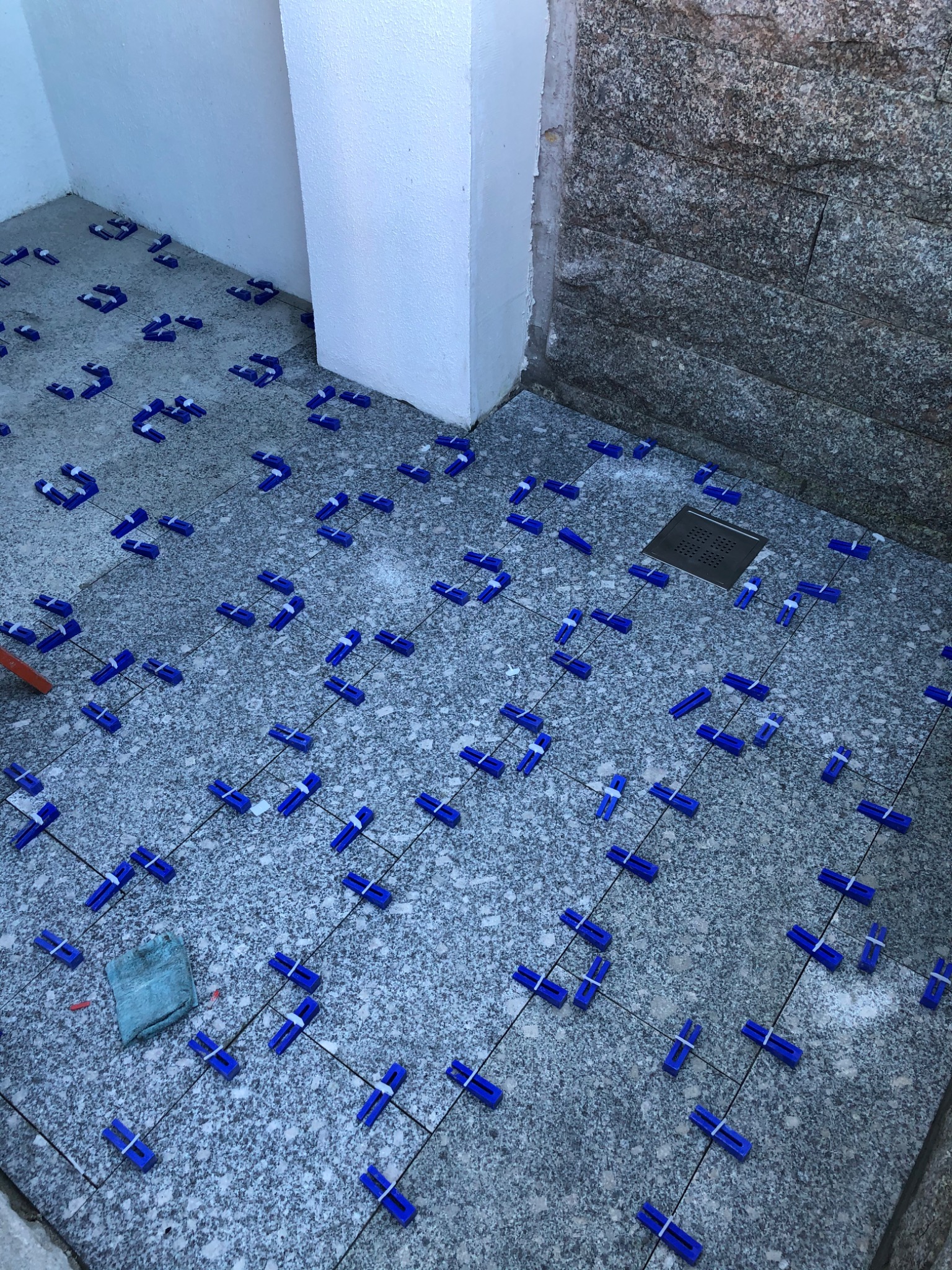 Granitowa posadzka w trakcie układania, z użyciem niebieskich klipsów poziomujących i sznurka, widoczny odpływ liniowy i szmatka do czyszczenia.