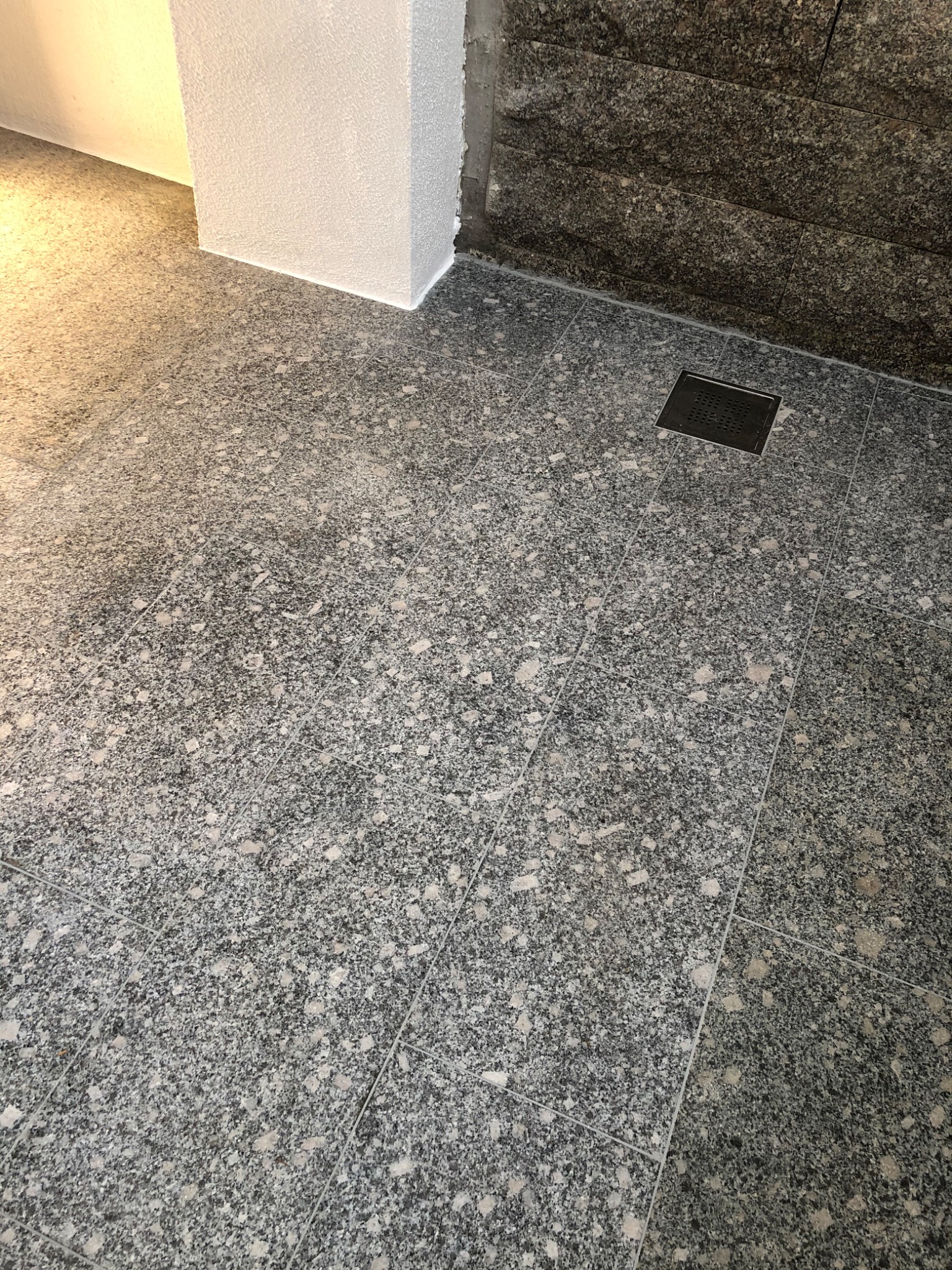 Szara, nakrapiana podłoga z płytek ceramicznych, z odpływem liniowym, przylegająca do ściany wyłożonej kamieniem dekoracyjnym i wykończonego narożnika ściany.