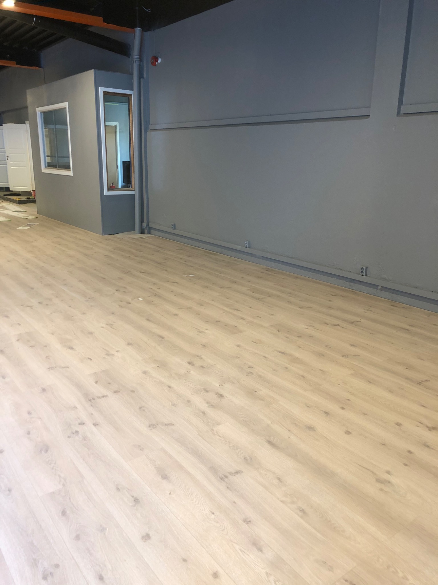 Jasne panele podłogowe ułożone w nowym, szarym pomieszczeniu z widoczną instalacją elektryczną przy ścianie i fragmentem kabiny biurowej.